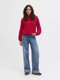 Röd stickad tröja med ett strukturerat mönster, ihop med blå vida jeans med en vikt kant och bruna skor.