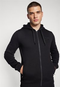 Felpa nera con zip realizzata in tessuto morbido, dotata di chiusura frontale con cerniera, cappuccio con cordino e tasche laterali. Piccola patch con logo sul petto.