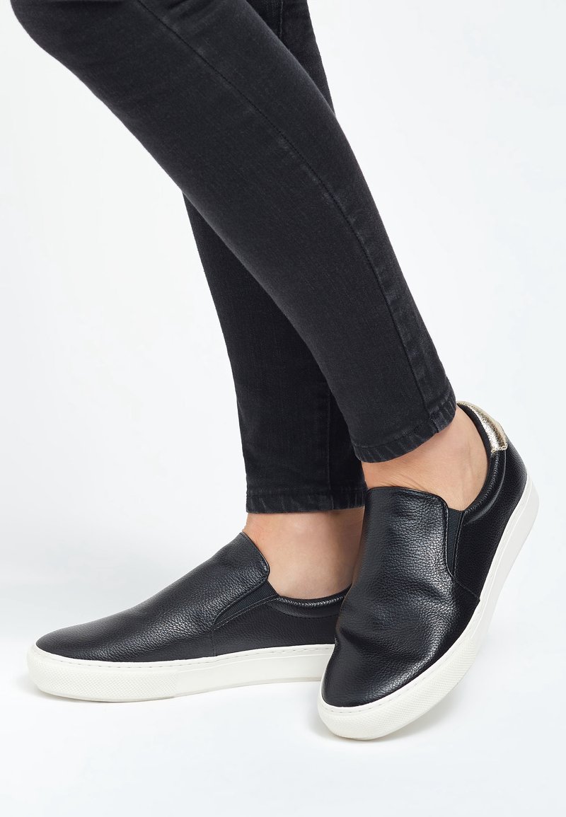 Personne portant des baskets en cuir noires sans lacets avec des semelles blanches et un jean skinny noir, debout sur une surface blanche.