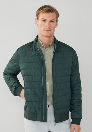 Chaqueta acolchada de color verde oscuro con cremallera en el frente y puños acanalados. Llevada sobre una camisa verde claro, mostrando un look casual en capas.