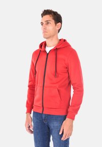 Felpa rossa con zip e tasca a marsupio, cordini neri e polsini a coste. Abbinata a jeans blu, con una maglietta bianca semplice sotto.