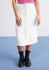 Biely denim midi sukňa s prednými gombíkmi, mierne rozšírený dolný okraj, nosená s čiernymi šnurovacími bojovými topánkami so žltým prešívaním.