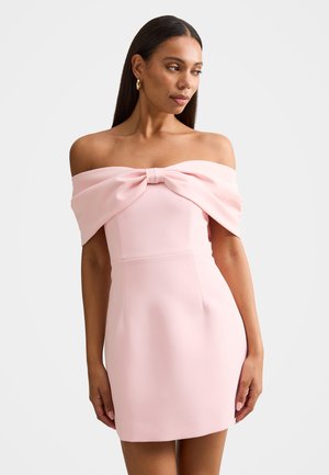 Žena s dlhými tmavými vlasmi, oblečená v svetloružových šatách s odhalenými ramenami a veľkou mašľou na výstrihu, stojaca na bielom pozadí.