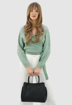 Frau mit langen, welligen Haaren, die einen grünen Wickelpullover und weiße Hose trägt und eine schwarze Ledertasche an den Griffen hält.