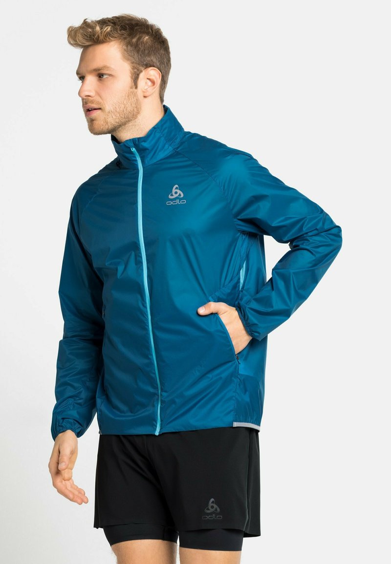 Türkisfarbene leichte Jacke mit Stehkragen, Frontreißverschluss und elastischen Bündchen. Mit einem Logo auf der Brust und seitlichen Taschen. Getragen mit schwarzen Shorts.