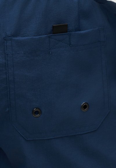 Τσέπη από ύφασμα ναυτικού μπλε με μαύρη ετικέτα από πάνω και δύο μεταλλικά φωλιά κοντά στο κάτω κέντρο, ραμμένη με ενισχυμένα ραφές.