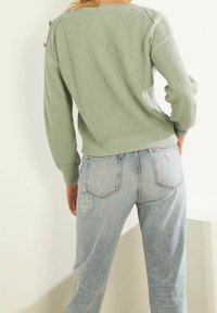 Light green ribbed knit sweater with a round neckline, featuring button accents on the shoulder, paired with stylish light blue distressed jeans.