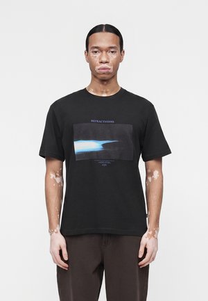 Vitiligós férfi fekete mintás pólót és sötét nadrágot visel, egy egyszínű világos háttér előtt áll.