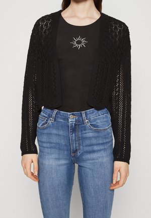 Svart öppenstickad cardigan med avslappnad passform, med långa ärmar. Bärs över en svart topp med en silverfärgad solstråle-design, matchad med blå jeans.
