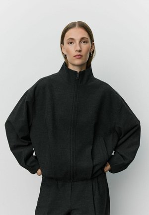 Kobieta o prostych włosach nosi ciemnoszarą kurtkę zapinaną na zamek z wysokim kołnierzem i pasujące spodnie, stoi z rękami w kieszeniach na jednolitym tle.