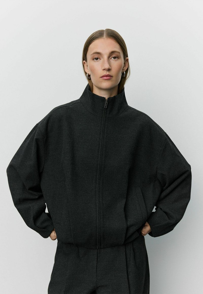 Femme aux cheveux raides portant une veste zippée gris foncé à col montant et un pantalon assorti, debout les mains dans les poches devant un fond uni.