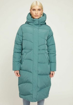 Woman wearing a long teal puffer coat with a high collar and hood, standing against a plain light background.