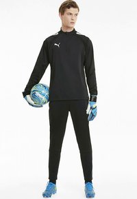 Fiatal férfi labdarúgó kapus fekete Puma öltözetben és kék kesztyűvel tartja a kék és zöld Puma labdát.