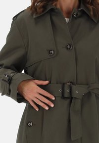 Cappotto trench verde oliva caratterizzato da un design a doppio petto, grandi bottoni, vita con cintura e polsini regolabili con eleganti dettagli metallici.