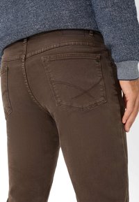 Jeans di denim marrone a vestibilità slim, con tasche posteriori e dettagli di cucitura discreti. Indossati con un maglione blu navy dalla texture.