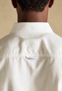 Vue de dos d'une personne portant une chemise blanche à col avec une petite étiquette "JOULES" cousue sous le col.