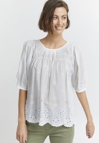 Blouse en coton blanc avec des manches bouffantes, présentant des broderies florales, un ourlet festonné et des détails en œillet le long du bord inférieur.
