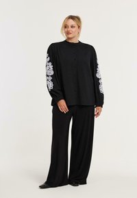 Schwarze Bluse mit weißen gestickten Mustern an den Ärmeln, mit Knopfleiste, hohem Kragen und lockerer Passform von schwarzen Hosen.