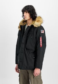 Jachetă parka neagră cu glugă din blană artificială, multiple buzunare și un emblema pe braț. Prezintă un fermoar cu detaliu roșu.