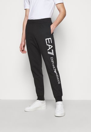 Person trägt schwarze Jogginghosen mit weißem EA7 Emporio Armani-Logo am linken Bein, kombiniert mit weißen Sneakers und einem weißen T-Shirt.