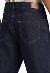 Mörkblå denimjeans med kontrasterande orange sömmar, två bakfickor och en brun läderlapp med en logotyp på midjebandet.