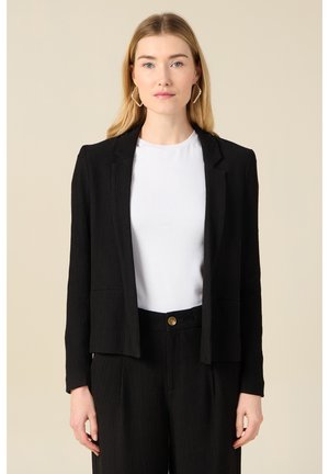 Blazer noir texturé avec un col, des poches avant et un design court. Porté sur un t-shirt blanc, associé à un pantalon noir.