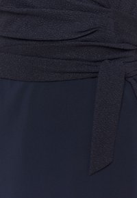 Námořnické šaty s řasenou, třpytivou horní částí a hladkou, matnou sukní. Obsahují detail vázacího pásku v oblasti pasu a texturovaný povrch.