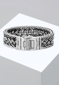 Silverfärgat metalarmband med vävd design, med en rektangulär låsning där "Kuzoi" är graverat. Slät yta, polerad finish.