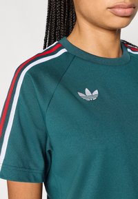 Zöld sportos póló kerek nyakkivágással, fehér és piros csíkokkal a vállakon. Az Adidas logó szürke színben a mellrészen.