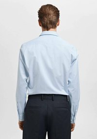 Camicia a maniche lunghe azzurro chiaro con sottili strisce, dotata di colletto classico e polsini con bottoni, abbinata a pantaloni scuri, vista da dietro.