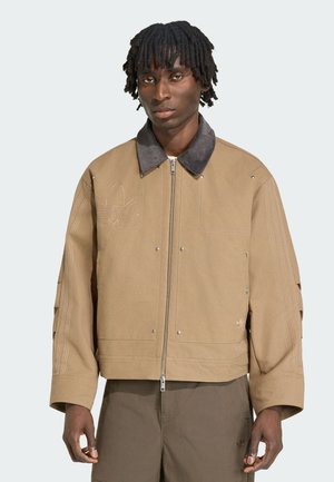 Ung mand med mørk hud og dreadlocks iført en beige lynlåstrøje med mørkrørt krave og matchende brune bukser mod en ensfarvet baggrund.