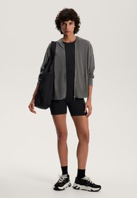 Zwarte fietsshorts, grijze hoodie met ritssluiting, zwarte tote bag. Model draagt zwarte sneakers met witte accenten, korte krullende haar, neutrale achtergrond.