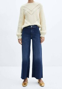 Roomkleurige gebreide trui met een chevronpatroon, gecombineerd met donkerblauwe wijde jeans en gouden instapschoenen. De outfit is casual en textuurrijk.