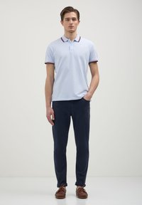 Uomo in piedi con una mano in tasca, indossando una polo azzurra chiara, pantaloni blu scuro e scarpe marroni con lacci, su uno sfondo semplice.