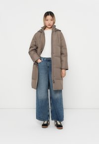 Manteau long rembourré marron avec une capuche, fermeture éclair à l'avant et poches latérales. Porté sur un haut blanc et un jean bleu à jambes larges.