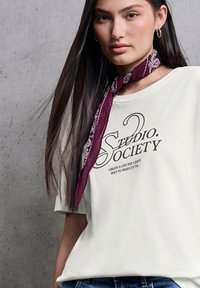 Weißes T-Shirt mit kurzen Ärmeln und einem braunen Drucklogo "Studio Society" sowie dem Text darunter. Das Model trägt einen burgunderfarbenen gemusterten Schal um den Hals.