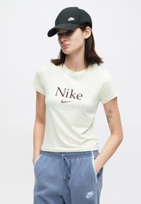 Mladá žena v bielej tričku Nike, modrých teplákových nohaviciach Nike a čiernej čiapke Nike, stojí s rukami v vreckách a neutrálnym výrazom.