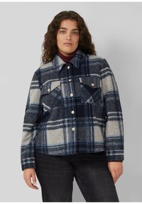 Frau mit lockigem Haar, die eine blau-grau karierte Jacke über einem weinroten Rollkragenpullover und schwarze Hosen trägt, stehend vor einem grauen Hintergrund.