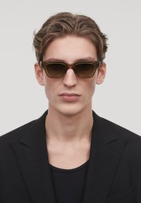 Lunettes de soleil en acétate marron avec des montures rectangulaires, des verres teintés sombres et des accents en métal. Modèle portant un blazer noir sur un top noir.