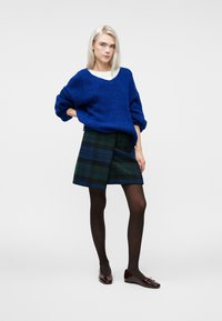 Niebieski sweter o grubym splocie z dekoltem w kształcie litery V, w połączeniu z ciemną spódnicą w kratę. Modelka nosi czarne rajstopy i błyszczące burgundowe baleriny.