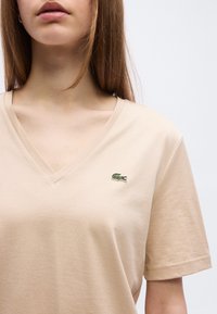 Γυναίκα που φοράει μια μπεζ μπλούζα με λαιμόκοψη V και μικρό πράσινο λογότυπο κροκοδείλου κεντημένο στο αριστερό στήθος.