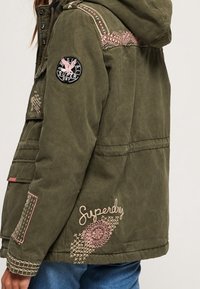 Olijfgroene jas met capuchon, geborduurde patronen in het roze, een zwarte cirkelvormige logopatch op de mouw en details van textuurstof.