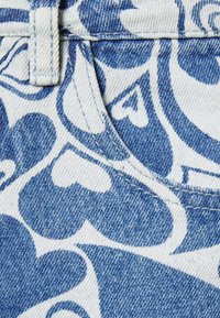 Tecido de denim com um padrão de corações brancos e azuis, com costuras visíveis ao longo das costuras e da área dos bolsos. Acabamento texturizado.