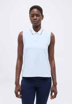 Μαύρη γυναίκα που στέκεται, φοράει αμάνικο γαλάζιο πόλο μπλουζάκι με σκούρο μπλε παντελόνι, μπροστά σε απλό λευκό φόντο.