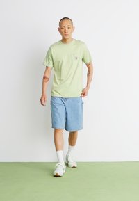 Lysegrønn kortermet t-skjorte med brystlomme, kombinert med lyseblå denimshorts. Modellen har på seg hvite joggesko og ankelsokker.