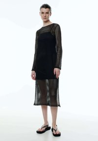 Schwarzes Mesh-Kleid mit langen Ärmeln, halbtransparenter Stoff und geradem Schnitt. Getragen mit schwarzen Flip-Flop-Sandalen.