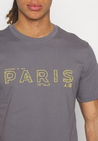 Muž v šedém tričku s žlutě orámovaným textem "PARIS" a malými logy a kódy pod písmeny.