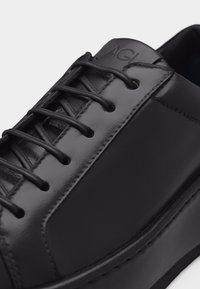 Baskets en cuir noir avec lacets noirs, panneaux lisses et texturés, coutures visibles et initiales de la marque "AGL" en relief sur la languette.