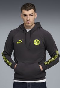 Ung mand iført en sort Puma hoodie med BVB 09-logo og gule mønstrede striber ned ad ærmerne, hænderne i forlommerne, neutral grå baggrund.