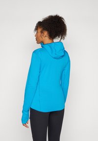 Blå hettejakke med teksturert stoff, tettsittende design og lange ermer. Har en litt buet kant og tommelhull i ermene.